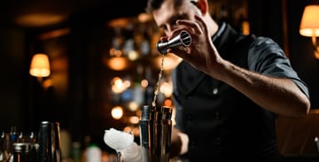 A flair bartender high-pours a shot into a shaker tin