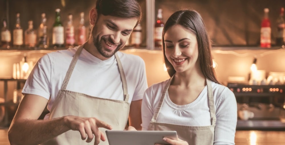 Two bartenders standing side by side, looking at a tablet.