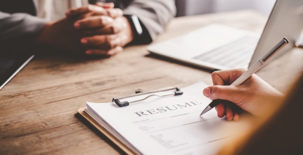 Person sitting at table with hands clasped, resume across the table being reviewed by another individual.